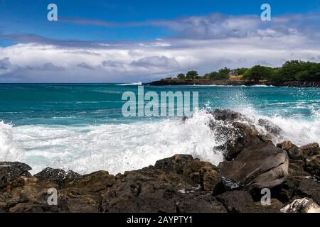 Welle bricht an rauem Ufer der Kona-Küste, auf der großen Insel von Hawaii. Blaugrüner Pazifischer Ozean jenseits; felsige Küste mit Bäumen in der Ferne. C Stockfoto