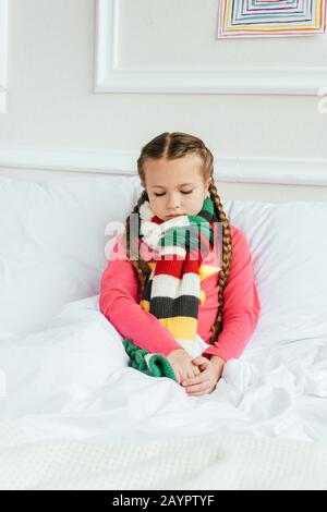 Trauriges krankes Kind im Schal, das auf dem Bett sitzt Stockfoto