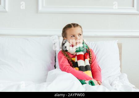 Trauriges krankes Kind im Schal, das auf dem Bett sitzt Stockfoto