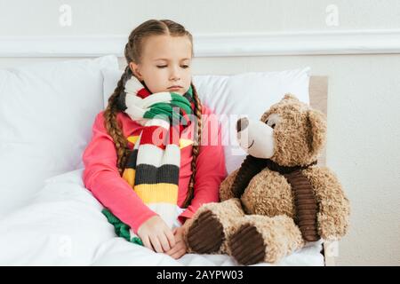 Trauriges krankes Kind im Schal, das mit Teddybär auf dem Bett sitzt Stockfoto