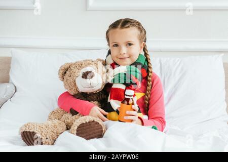 Lächelndes krankes Kind im Schal, das Sirup hält und mit Teddybär auf dem Bett sitzt Stockfoto