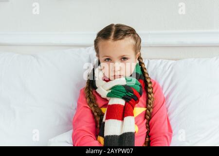 Trauriges krankes Kind im Schal, das auf dem Bett sitzt Stockfoto