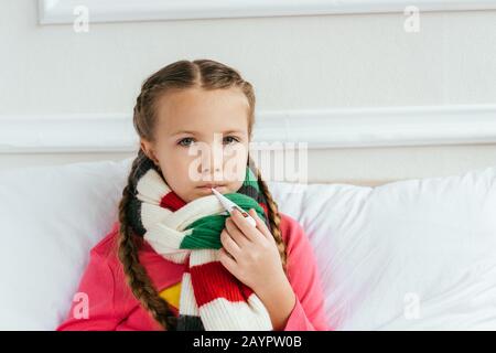 Trauriges krankes Kind im Schal, das Fieber hat und Thermometer auf dem Bett hält Stockfoto