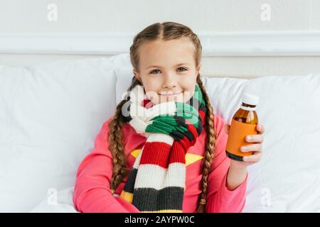 Lächelndes krankes Kind im Schal, der Sirup hält und auf dem Bett sitzt Stockfoto