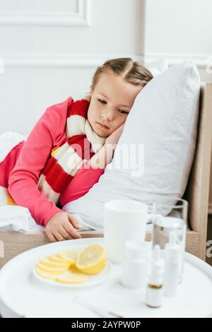 Trauriges krankes Kind im Schal, das auf dem Bett liegt und Medikamente in der Nähe hat Stockfoto