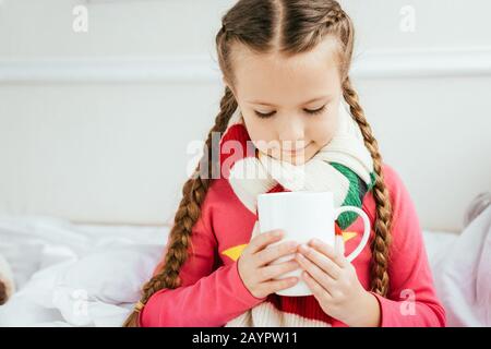 Süßes, krankes Kind im Schal, das eine Tasse heißen Drink hält, während es auf dem Bett sitzt Stockfoto