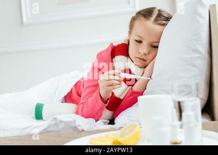 Krankes Kind mit Fieber im Schal, das auf Thermometer schaut, während es mit Medikamenten in der Nähe auf dem Bett liegt Stockfoto