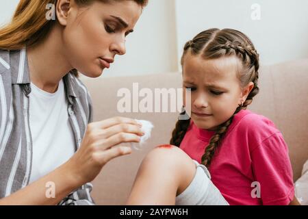 Mutter hält antiseptische Baumwolle in der Nähe der Wunde am Knie der traurigen Tochter Stockfoto