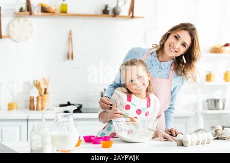 Selektive Fokussierung von Mutter und Tochter lächelnd und kochende Cupcakes mit Zutaten wie Eiern, Milch und Mehl Stockfoto