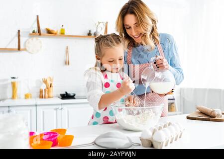 Selektiver Fokus der Tochter, die Teig knetet, während die Mutter zu Hause Milch aus der Kanne in die Schüssel gießt Stockfoto