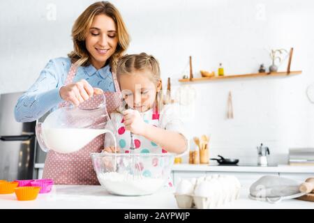 Tochter knetet Teig, während Mutter Milch aus Kanne in die Schüssel in der Küche gießt Stockfoto