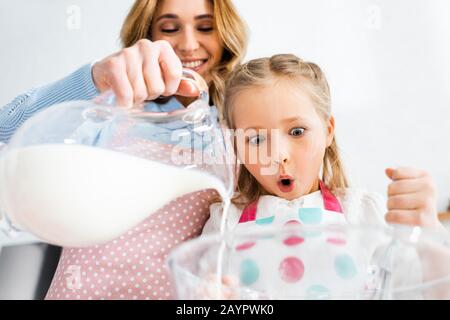 Niedriger Winkel der schockierten Tochter, die die Mutter betrachtet, die Milch aus der Kanne in die Schüssel gießt Stockfoto