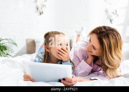 Lächelnde Mutter sieht schockierte Tochter mit digitalem Tablet im Schlafzimmer an Stockfoto