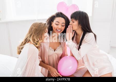 Fröhliche multikulturelle Freundinnen flüstern und klatschen im Bett mit pinkfarbenen Ballons Stockfoto