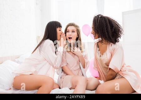 Schockierte multikulturelle Freundinnen flüsternden und klatschenden mit pinkfarbenen Ballons im Bett Stockfoto