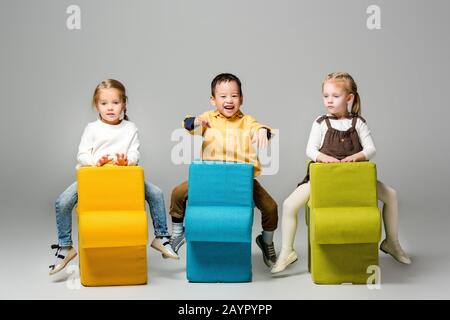 Fröhliche multikulturelle Kinder sitzen auf Rätselstühlen, auf Grau Stockfoto