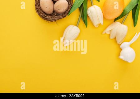 Draufsicht über Tulpen und Hühnereier im Gelege in der Nähe von Osterhase auf farbenfrohem gelbem Hintergrund Stockfoto