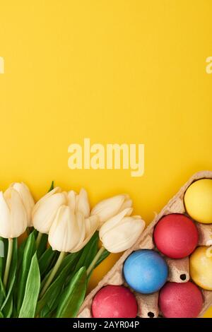 Draufsicht über Tulpen und mehrfarbig bemalte Ostereier in Pappkarton auf bunt gelbem Hintergrund Stockfoto