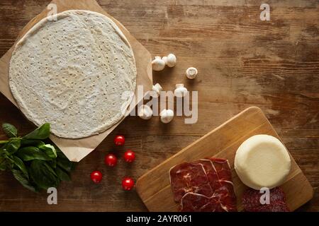 Draufsicht auf rohen Teig, Kirschtomaten, Basilikum, Pilze, Mozzarella, Salami und Prosciutto für Pizza auf Holztisch Stockfoto