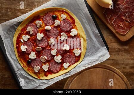 Draufsicht auf leckere Pizza mit Salami und Pilzen auf Pergamentpapier auf Holzhintergrund Stockfoto