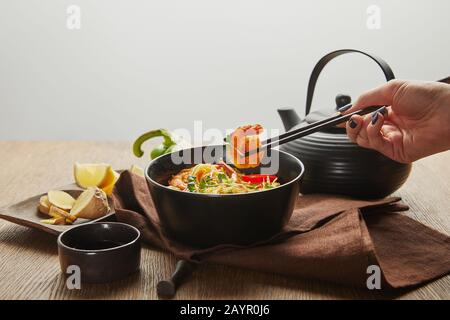 3/4-Ansicht der Frau, die Garnelen mit Schopfstäbchen in der Nähe von Nudeln in Schale hält, isoliert auf Grau Stockfoto