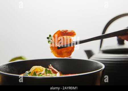 3/4-Ansicht der Frau, die Garnelen mit Schopfstäbchen in der Nähe von Nudeln in Schale hält, isoliert auf Grau Stockfoto