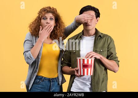 Verängstigte Mädchen, die Film ansehen, während sie mit einem Eimer Popcorn die Augen zu Freund schließen, isoliert auf Gelb Stockfoto