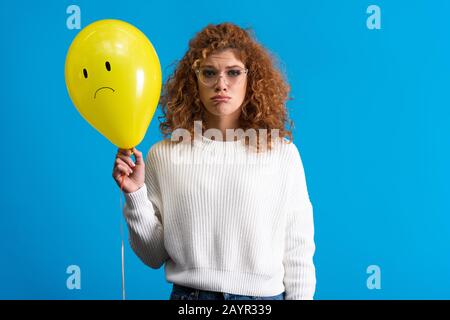 Trauriges Mädchen, das den gelben Ballon mit aufgebrachten Gesichts hält, isoliert auf Blau Stockfoto