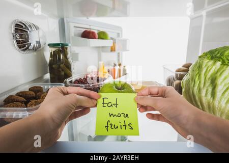 3/4-Ansicht der Frau, die Karte mit "Eat More Fruits"-Schriftzug mit Lebensmitteln im Kühlschrank auf weißem Stockbild abisoliert hält Stockfoto