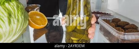 3/4-Ansicht der Frau, die einen Mixbecher mit Pickeln aus dem Kühlschrank mit Essen auf weißem Hintergrund, Panoramaaufnahme, Stockbild nimmt Stockfoto