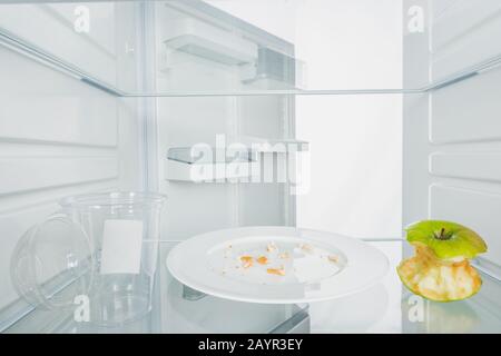 Kunststoffbecher mit gebissenen apfel und Krümel auf Platte im Kühlschrank mit offener Tür isoliert auf weißem, Stockbild Stockfoto