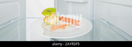 Panoramaaufnahme von gebissenen apfel mit Wurst und Krümel auf Platte im Kühlschrank mit offener Tür isoliert auf weißem, Stockbild Stockfoto