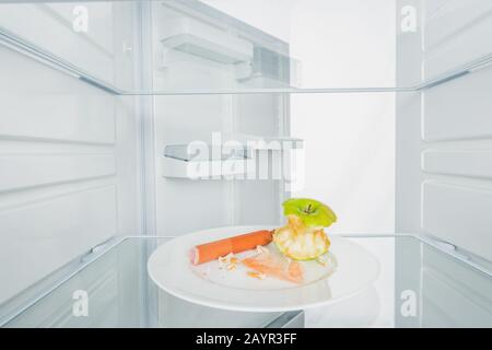 Nackter apfel mit Wurst und Krümel auf Platte im Kühlschrank mit offener Tür isoliert auf weißem, Stockbild Stockfoto