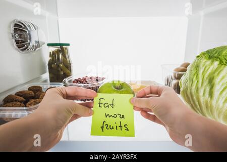 3/4-Ansicht der Frau, die Karte mit "Eat More Fruits"-Schriftzug mit Speisen im Kühlschrank im Hintergrund hält, isoliert auf weißem Stockbild Stockfoto