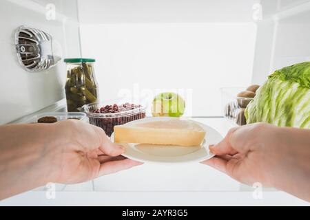 3/4-Ansicht der Frau, die Käse aus dem Kühlschrank nimmt, mit Früchten und Pickeln, isoliert auf weißem, Stockbild Stockfoto