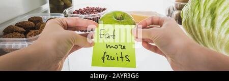 Panoramaaufnahme der Frau, die Karte mit "Eat More Fruits"-Schriftzug mit Speisen im Kühlschrank im Hintergrund hält, isoliert auf weißem Stockbild Stockfoto