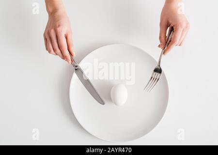 Draufsicht auf Frau, die Besteck in der Nähe von Ei auf weißem Hintergrund hält Stockfoto
