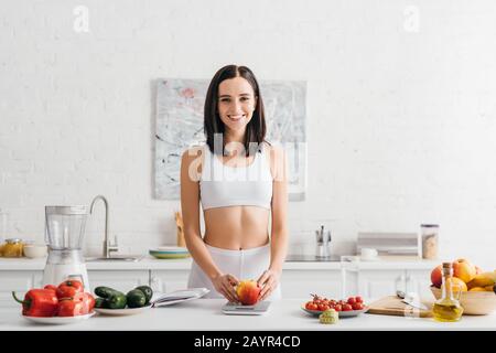 Lächelnde Sportlerin, die frischen apfel in der Nähe von Messband und Notizbuch auf Küchentisch wiegt, kalorienzählende Ernährung Stockfoto