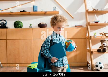 Intelligentes und lockiges Kind mit Rucksack, der Globus und Buch hält Stockfoto