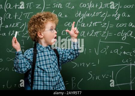 Überraschtes Kind in einer Brille, die mit mathematischen Formeln Kreide in der Nähe von Schwarzen Brett hält Stockfoto