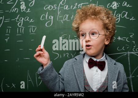 Smart Kind im Anzug und Bow Krawatte mit Kreide in der Nähe von Schwarzen Brett mit mathematischen Formeln Stockfoto