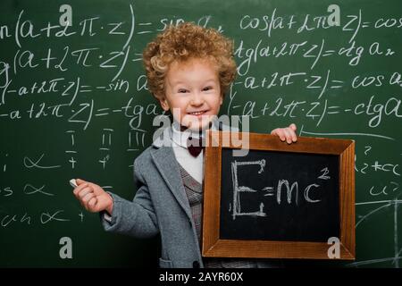 Lächelndes und intelligentes Kind im Anzug und mit Schleife, die kleine Tafel mit Formel in der Nähe von Schwarzen Brett hält Stockfoto