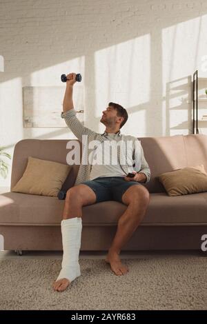 Gutaussehender Mann mit Bein im Pflasterbandtraining mit Hanteln und Fernbedienung auf dem Sofa Stockfoto