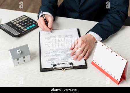 Freigestellte Ansicht des Realtor Signing Lease Agreement in der Nähe eines leeren Kalenders, eines Rechners und eines Hausmodells Stockfoto