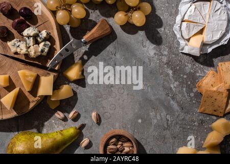 Draufsicht über Käse, Obst, Nüsse, Kräcker und Oliven mit Messer und Schneidebrett auf grauem Grund Stockfoto