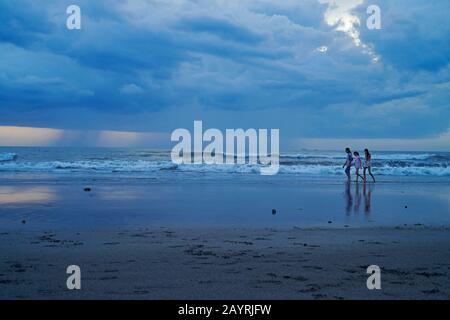 Mutter und ihre beiden Töchter gehen bei Sonnenuntergang Hand in Hand am Strand in Bali entlang Stockfoto