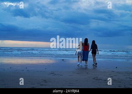 Mutter und ihre beiden Töchter gehen bei Sonnenuntergang Hand in Hand am Strand in Bali entlang Stockfoto