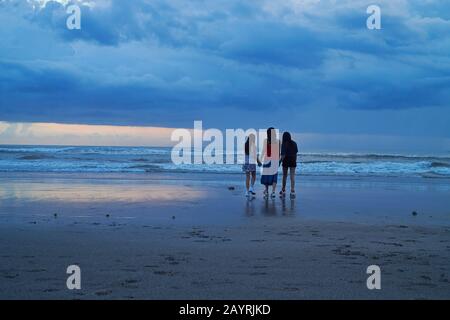 Mutter und ihre beiden Töchter gehen bei Sonnenuntergang Hand in Hand am Strand in Bali entlang Stockfoto