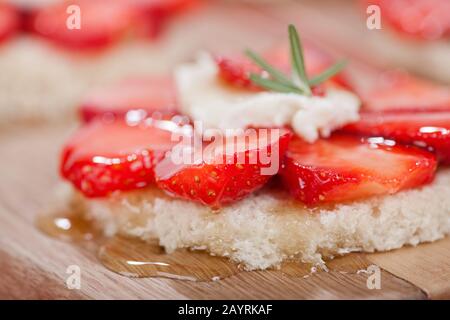 Geschnittene Erdbeeren auf Brot mit Ziegenkäse und Honig dröckeln darüber, mit einem Rosmarinzweig oben, der auf einem Holzschneidebrett ruht Stockfoto