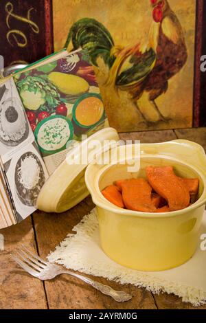 Gebackene kandierte Süßkartoffeln in einer antiken runden Kasserolenschale, die auf einer StoffServiette ruht, auf einer rustikalen Holztischplatte mit einem alten Kochbuch und Huhn Stockfoto